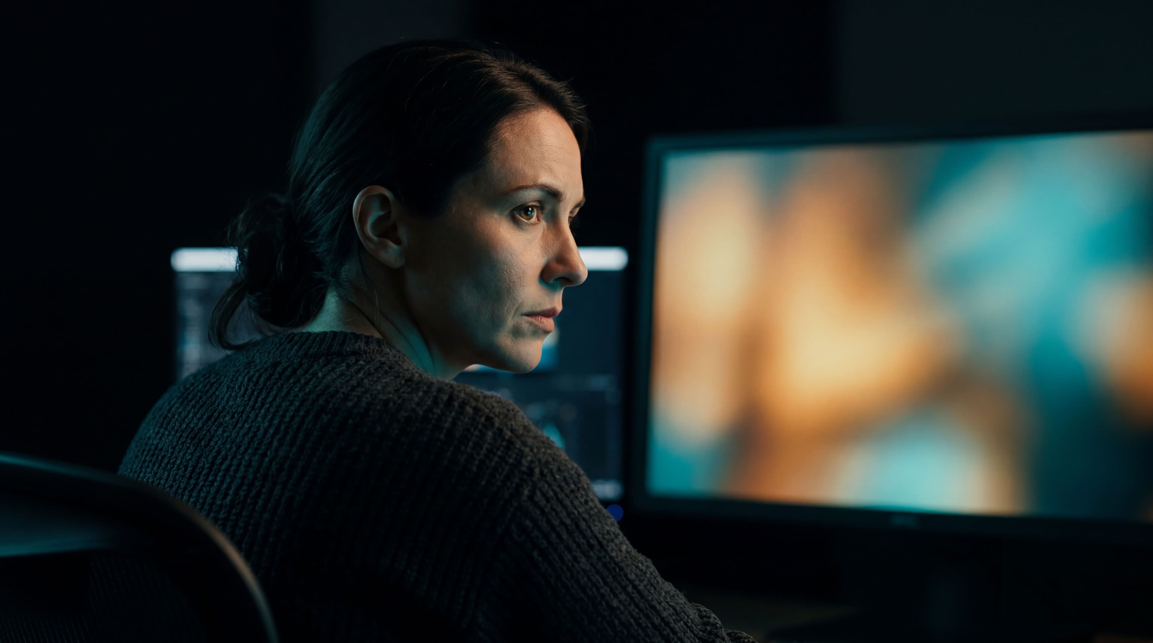 Profile of a focused woman in a darkened editing suite watching a reference monitor displaying a warm, slightly blurred cinematic frame, with secondary screens visible in the background.