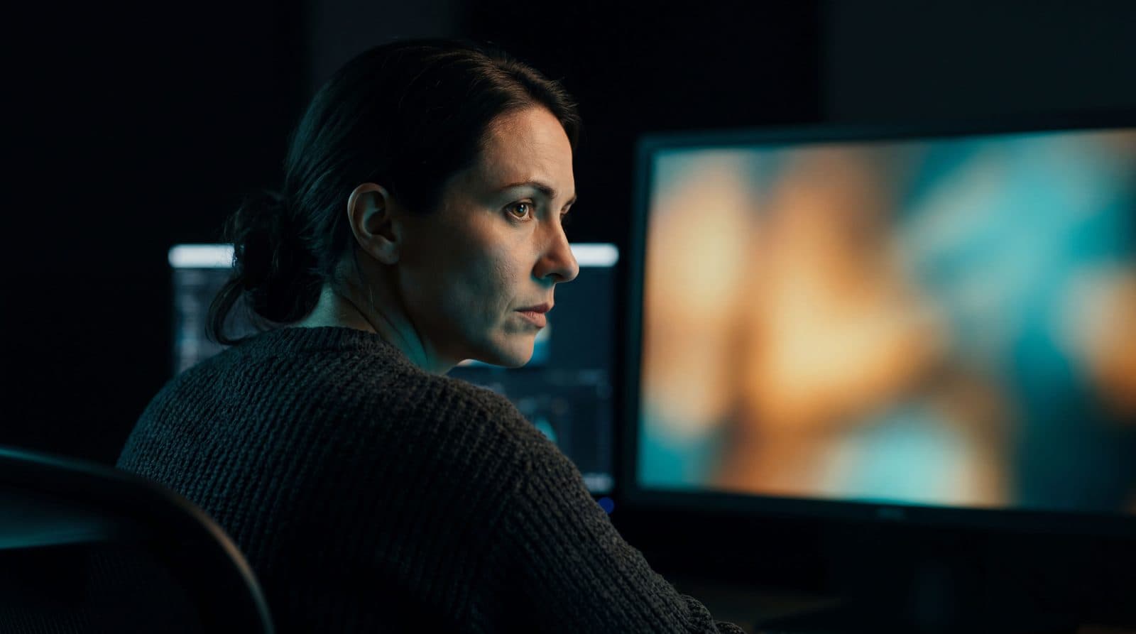 Profile of a focused woman in a darkened editing suite watching a reference monitor displaying a warm, slightly blurred cinematic frame, with secondary screens visible in the background.