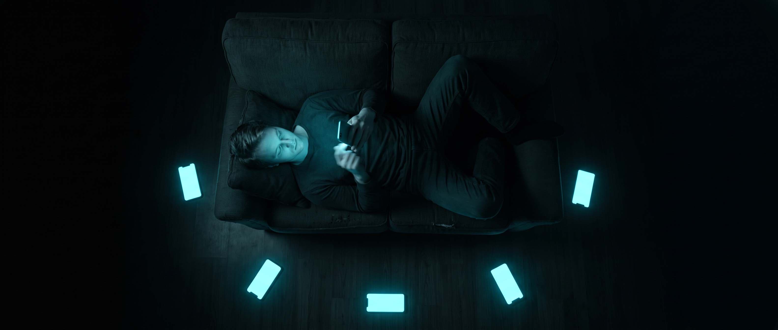Overhead view of a person lying on a couch in a dark room scrolling a phone, surrounded by several other glowing phone screens on the floor.