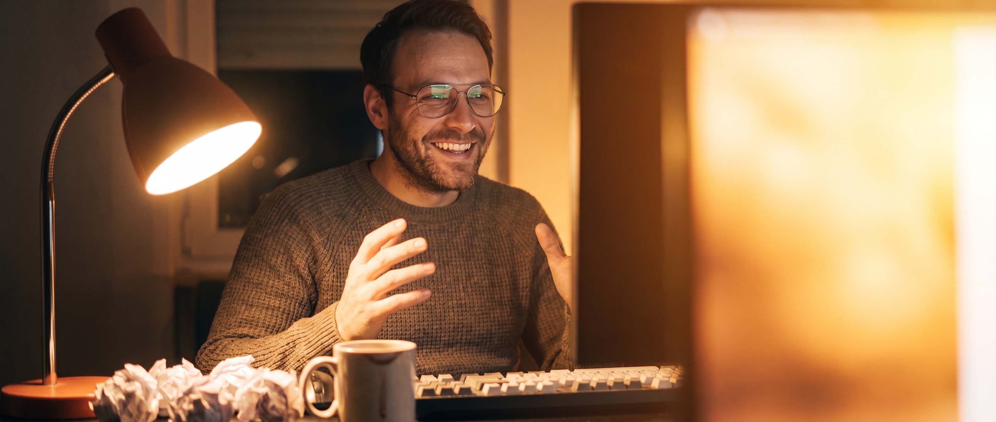 The same man smiling at his computer screen with both hands raised, lit by the warm glow of the monitor, crumpled paper still on the desk.