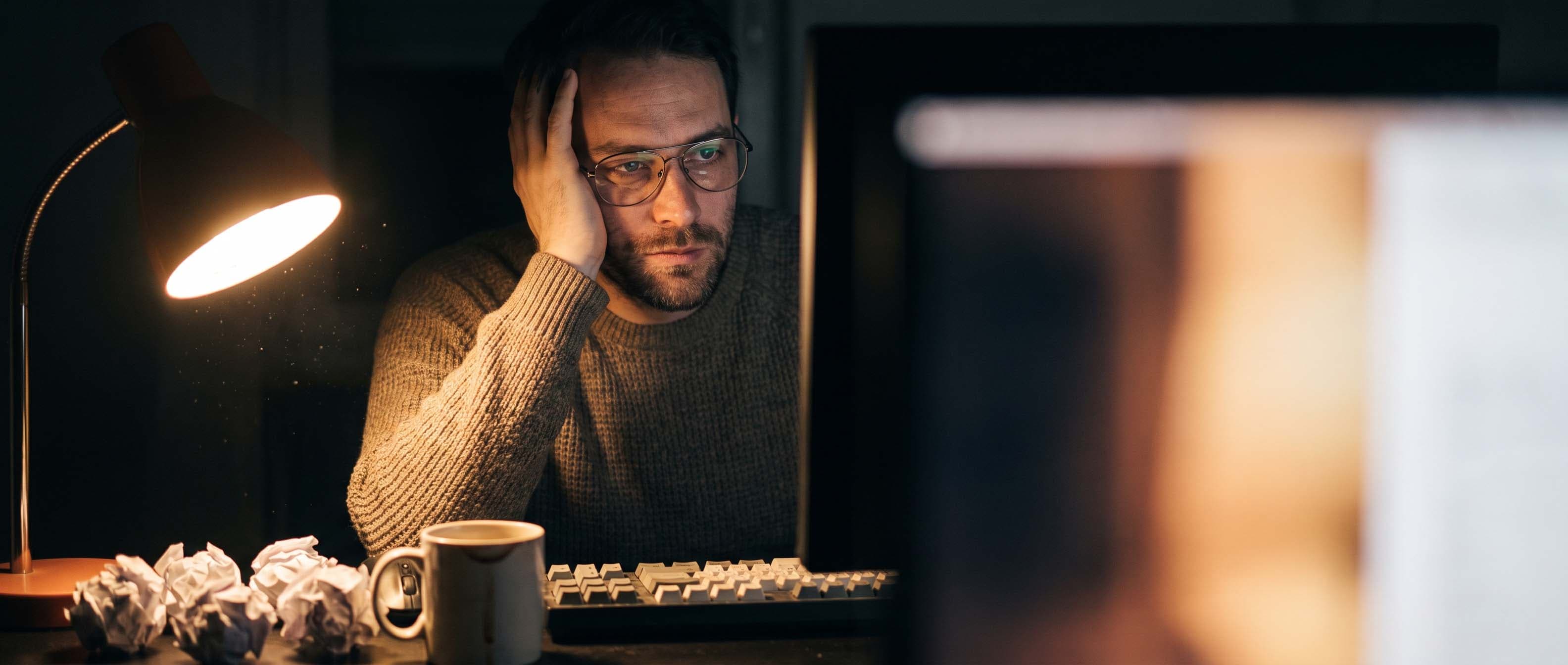 Man working late at a desk, head resting in his hand, staring at a computer monitor beside a lit desk lamp, a coffee mug, and crumpled sheets of paper.